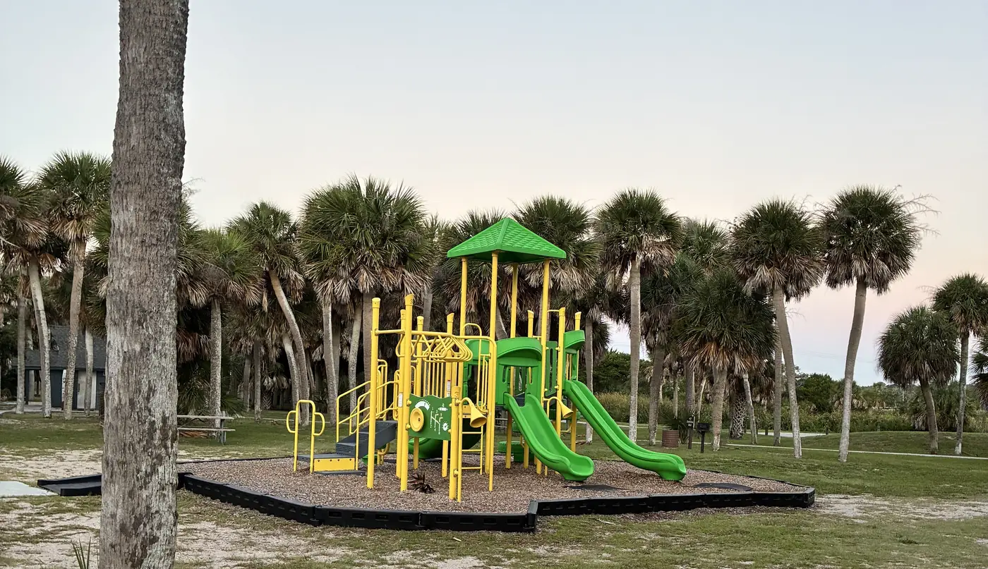 An ordinary green and yellow play structure on mulch, ringed by cabbage palms at dusk